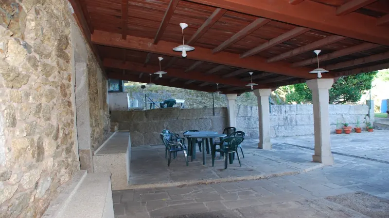 Zona de desayunos de Casa do Vento en Costa da Morte, situada en una terraza cubierta con techo de madera, columnas de piedra y mesas con sillas.